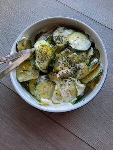 Schwedischer Gurkensalat mit Sahne in einer Schale mit Vorlegebesteck. Die Schale mit Gurkensalat steht auf einem Holztisch.