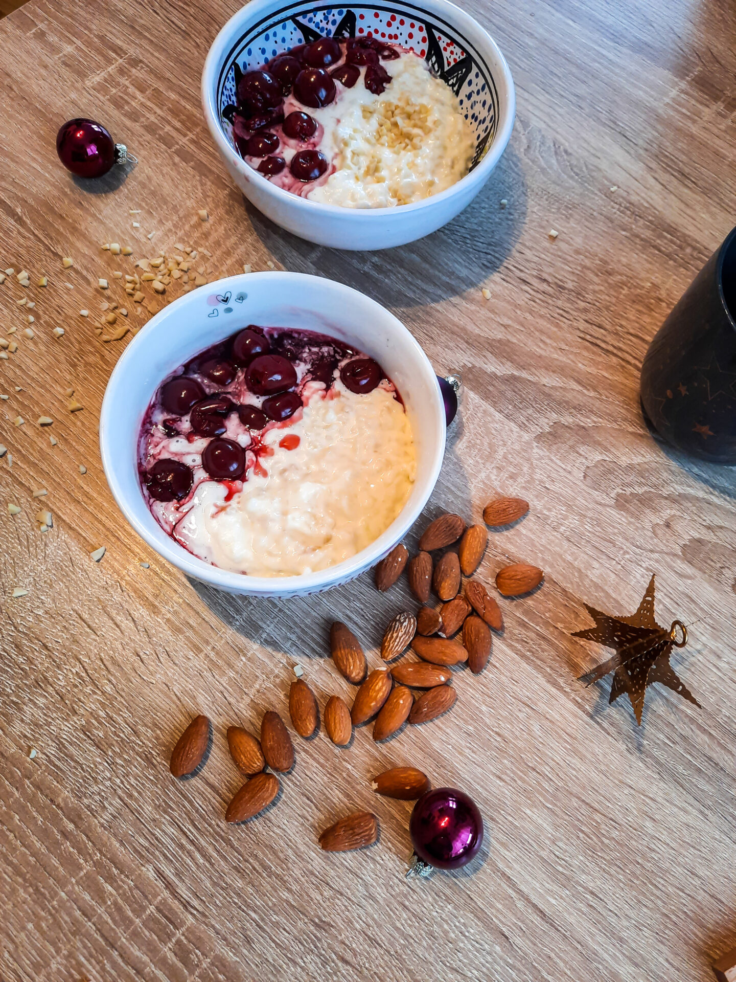 Zwei Schüsseln die gefüllt sind mit Milchreis mit Kirschen. Im Hintergrund liegen gehackte Mandeln, ganze Mandeln, zwei kleine Christbaumkugeln und ein Stern.