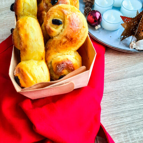 ein Körbchen mit Lussekatter, schwedischen Hefeteigschnecken, auf einem roten Tuch und eine Schale mit Weihnachtskugeln und Teelichern im Hintergrund.