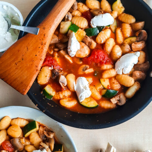 Eine Pfanne mit Gnocchi, Champignons und Zucchini steht im mittelpunkt. Auf ihrem Rand liegt ein Holzlöffel uund um Hintergrund liegen Champignons und Ricotta.