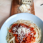 Teller mit Spaghetti und Linsenbolognese. Darauf geriebener Käse und im Hintergrund eine Käsereibe.