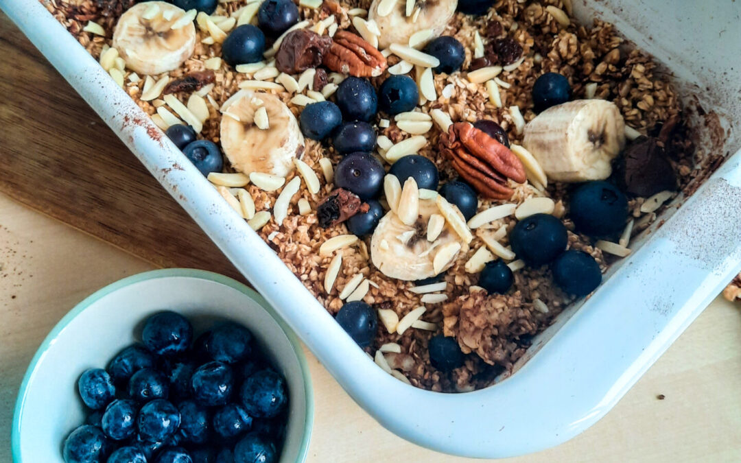 Eine Auflaufform mit Baked Oats ohne Banane, getoppt mit Banane, Blaubeeren und Mandeln. Davor eine kleine Schale mit Blaubeeren.