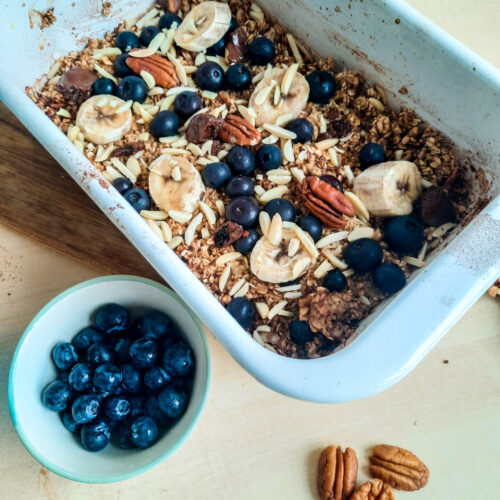 Eine Auflaufform mit Baked Oats ohne Banane, getoppt mit Banane, Blaubeeren und Mandeln. Davor eine kleine Schale mit Blaubeeren.