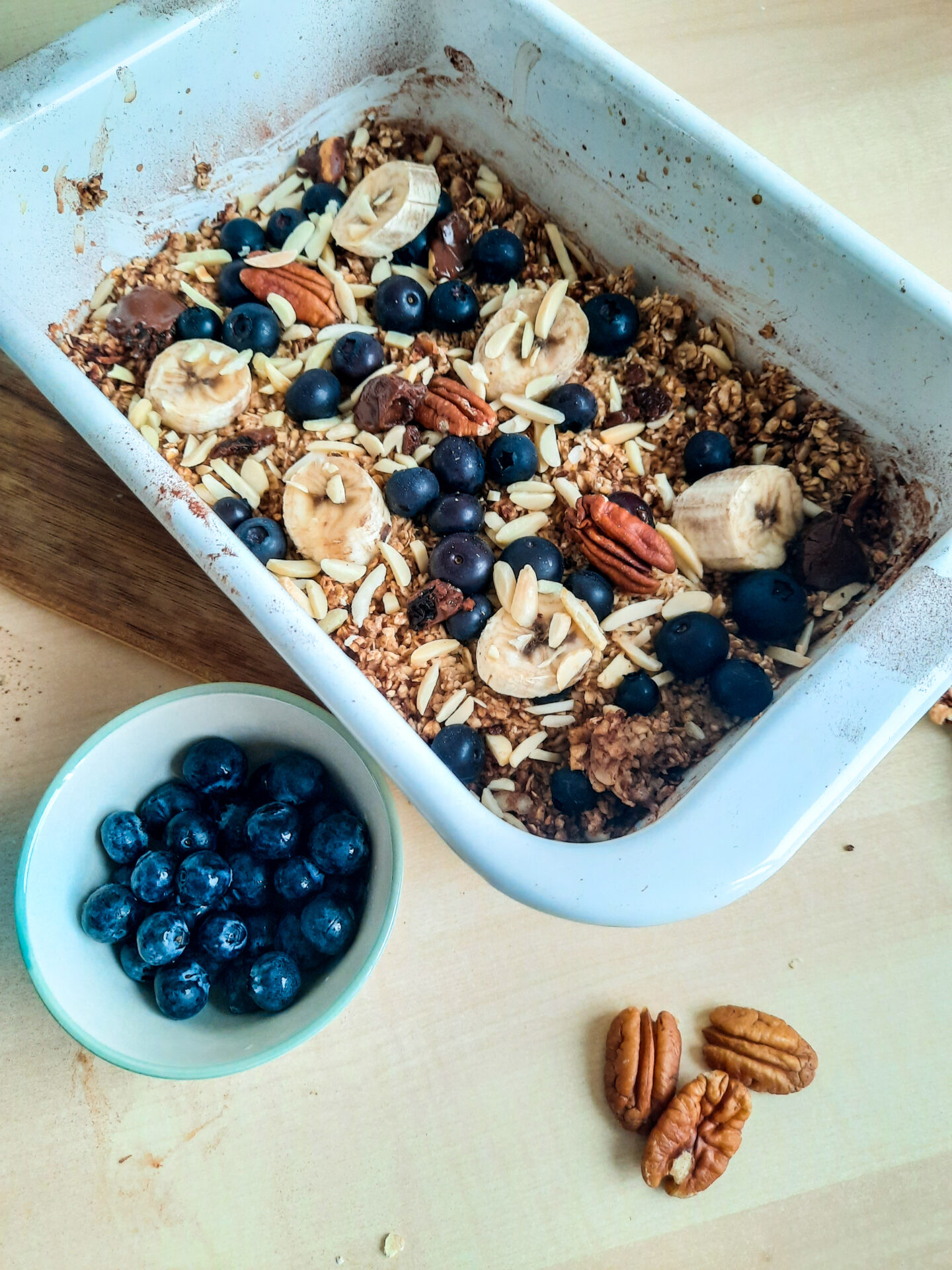 Eine Auflaufform mit Baked Oats ohne Banane, getoppt mit Banane, Blaubeeren und Mandeln. Davor eine kleine Schale mit Blaubeeren.