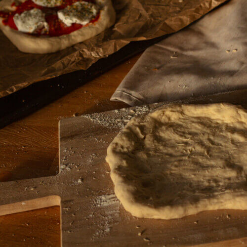 Vorne im Bild sieht man einen gerade ausgerollten Pizzateig mit Trockenhefe, der darauf wartet belegt zu erden. Daneben sieht man eine fertig belegte Pizza mit Tomaten und Mozarella auf einem Backblech.